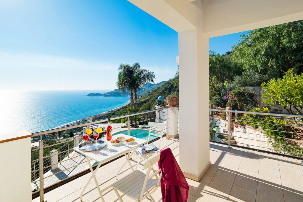 view from the balcony of Villa Kira, a villa in Taormmina. There's a table with snacks, the greenery and the sea view.