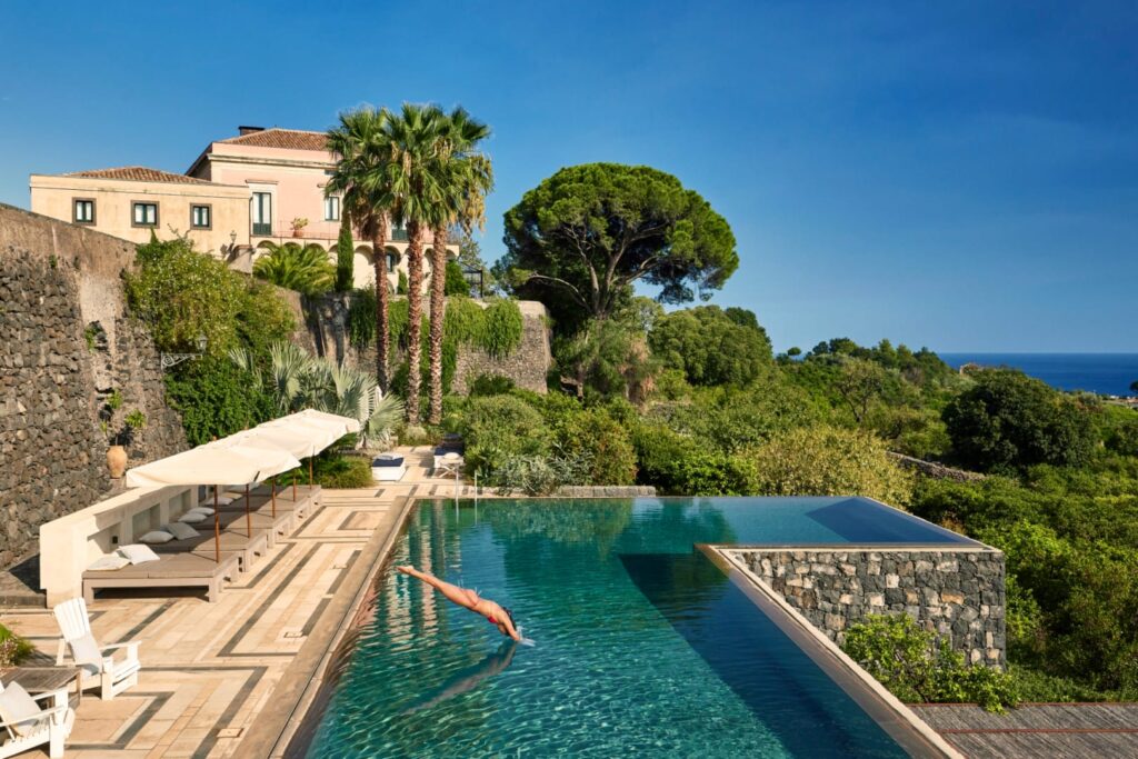 Private infinity pool of a villa in Sicily, Italy. In the foreground is a women diving in and sun beds across the outer edge of the pool. In the background there are various trees and other flora with the villa at the top of the hill.