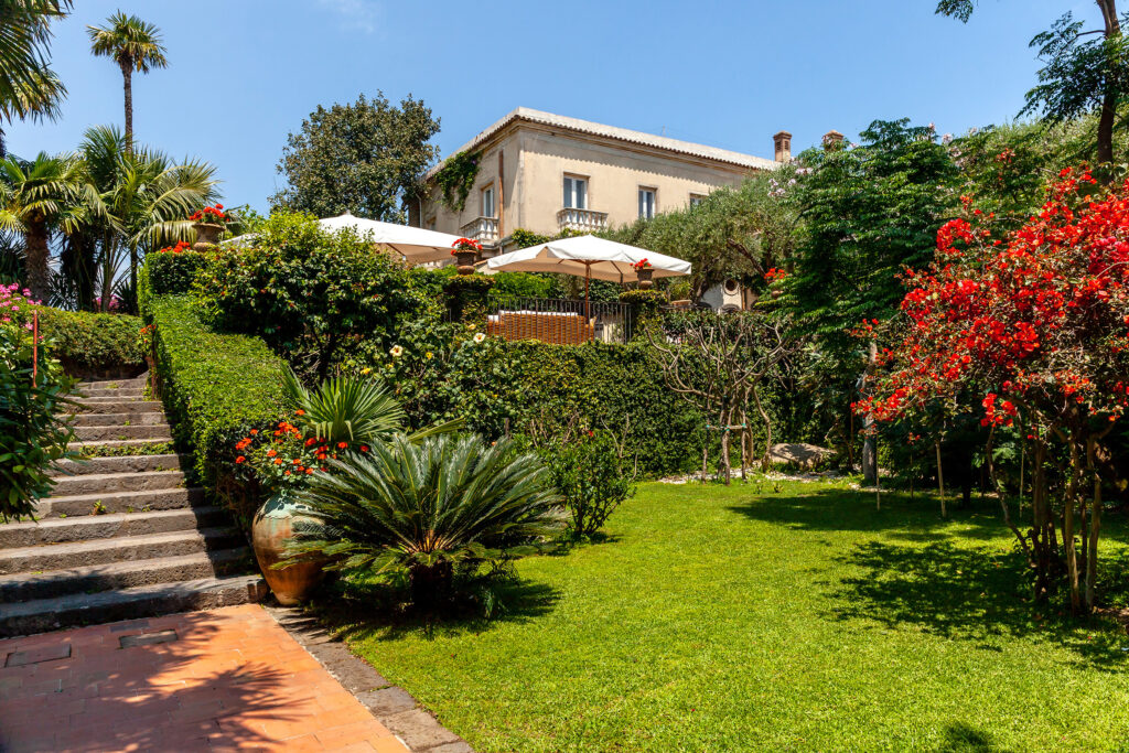 Private garden of a villa in Sicily. The foreground is the garden with lots of trees and red plants. There are stone stairs on the left leading up to the villa in the background.