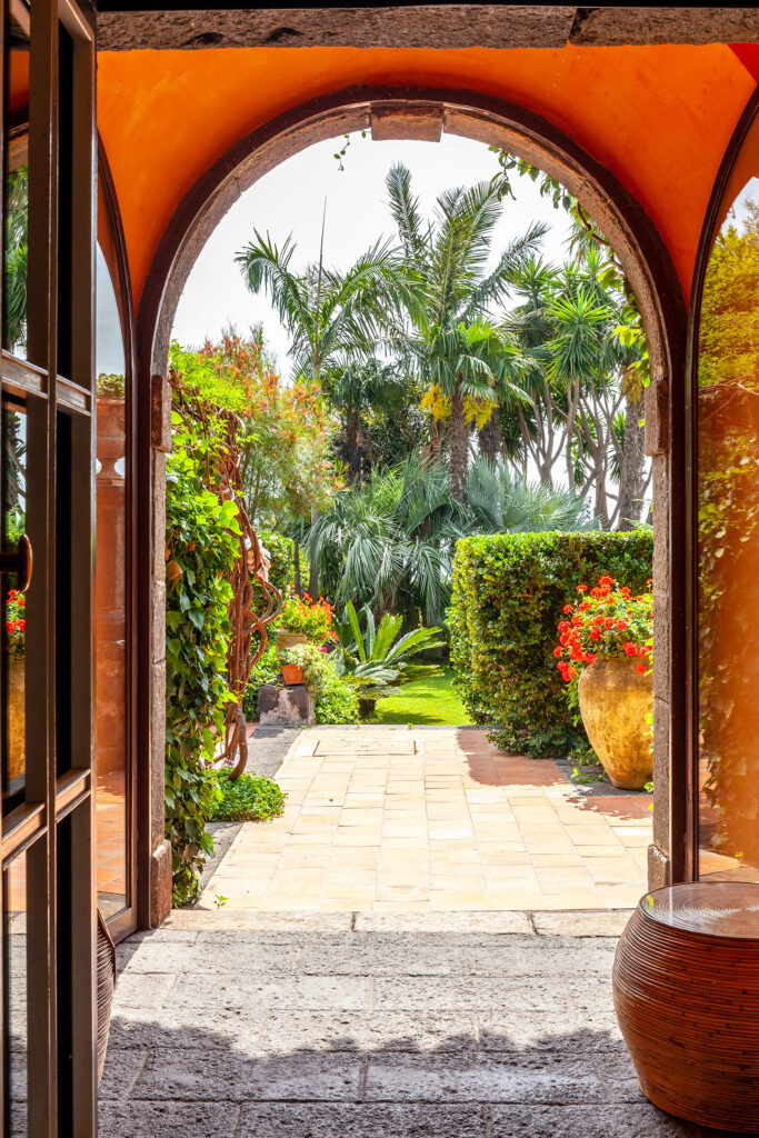 Doorway of a Sicilian Villa. The doorframe is wooden and the door itself is wood and glass, through the frame there is a stone path leading to the garden and fruit trees.