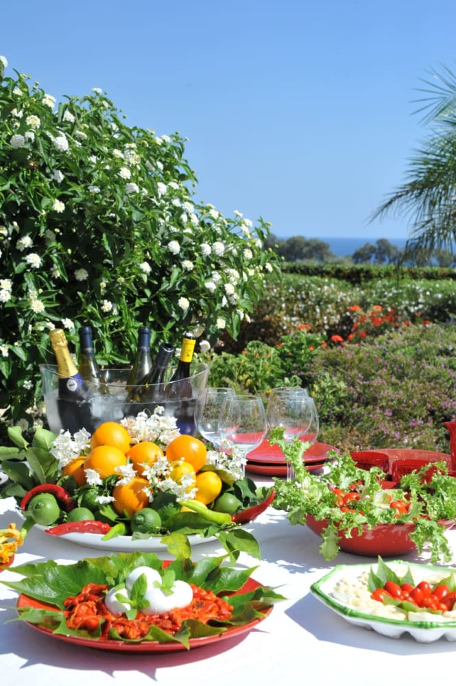 A table on the lemon garden premises with plates of food on white table cloth and a plate of oranges and lemons in the middle with wine behind.