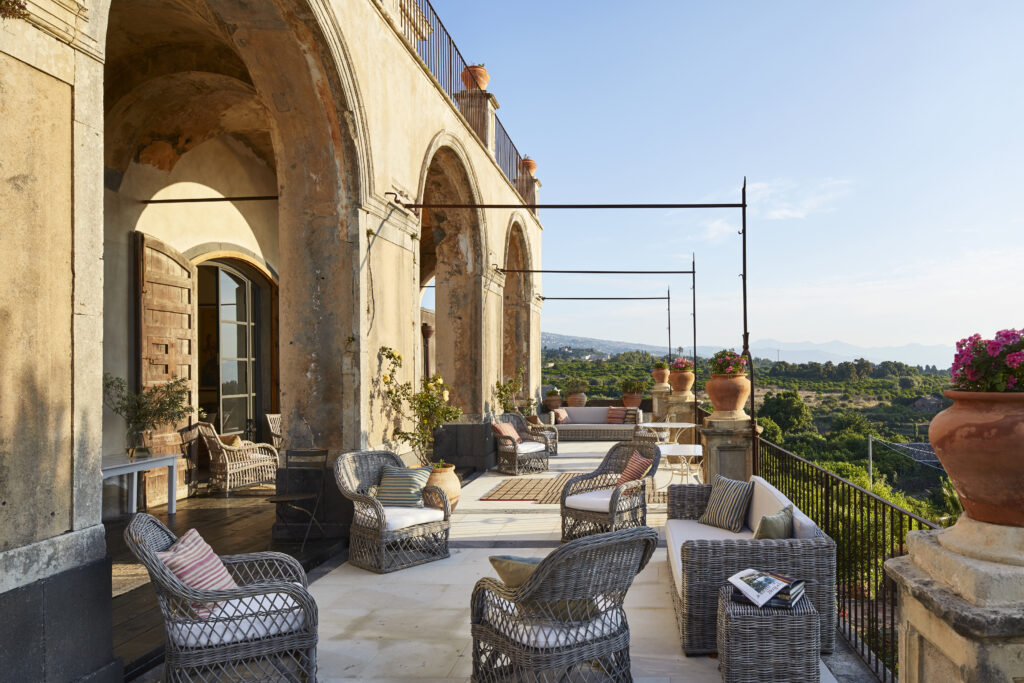Outdoor space of a villa in Sicily. There is a balcony/outdoor space made of stone, plants, and wicker chairs.