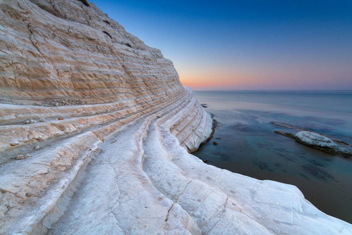 The rocky white cliffs of scala dei turchi at sunset. to the left is the white and orange cliff face, in the centre you see the sun setting and on the right is the deep blue sea.